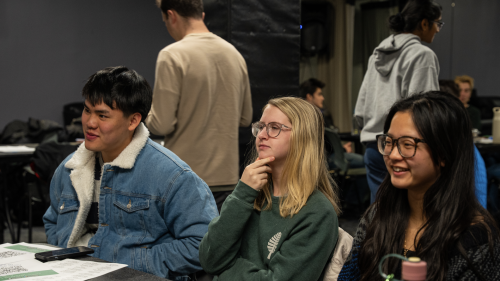 Three students sit at a table. They are listening to someone speak off camera.