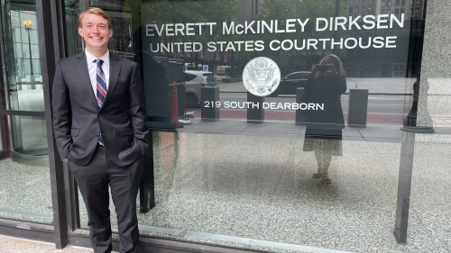 JJ Dega stands outside in a suit in front of a sign for the Everett McKinley Dirkesen Courthouse.