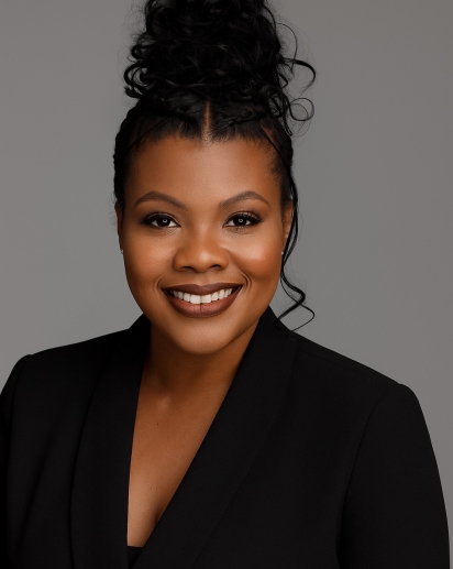 A headshot of Britni Jackson. She is wearing a black suit coat and is standing against a grey background.