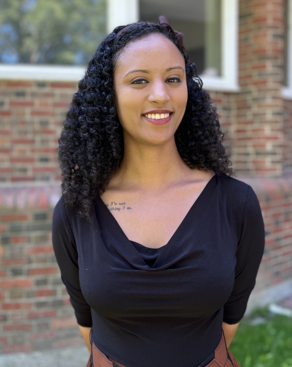 Tesha Davis standing outside against a brick wall. She is wearing a black top and an orange skirt.