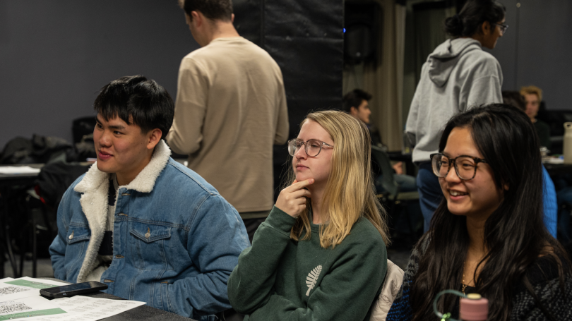 Three students sit at a table. They are listening to someone speak off camera.
