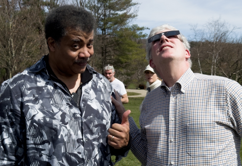 Neil deGrasse Tyson stands outside with a thumbs up next to Michael Rockefeller who is wearing black eclipse glasses and looking up into the sun.