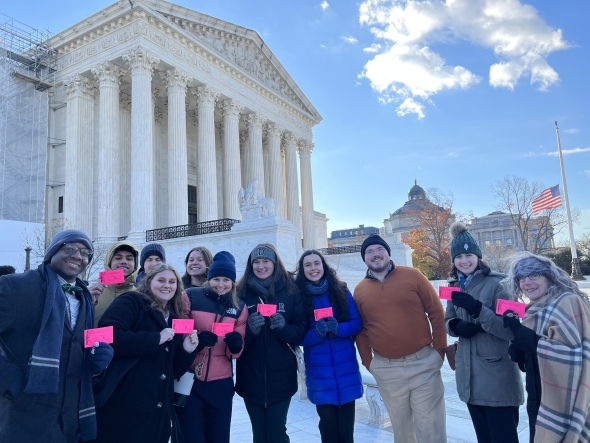 A group of students stand in front of the Supreme Court. They are holding pink slips.