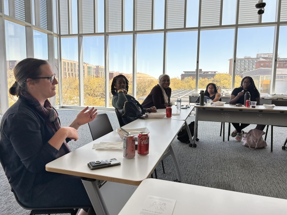 A group of scholars sit a round several tables in a conference room with large windows.